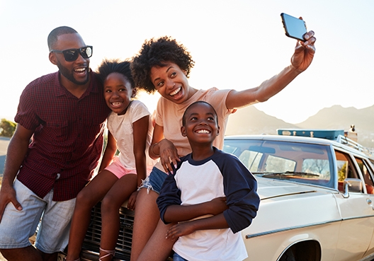 family of four sitting on, and others leaning on the bonnet of their car taking a selfie