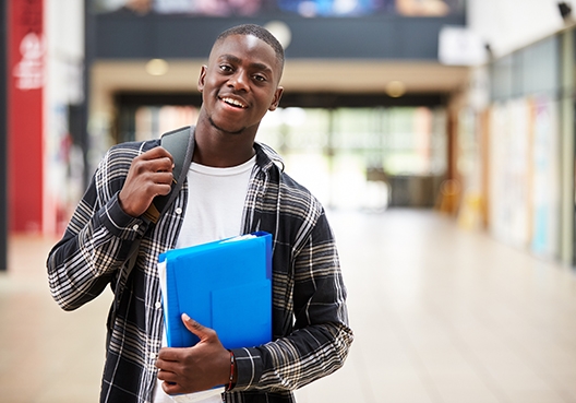 Student walking in the hallway of his school with a backpack over his shoulder and a folder in his arms