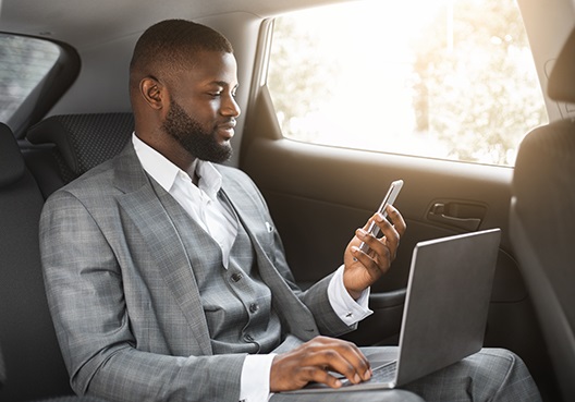 Guy sitting in the backseat of the car with his laptop, open on his laptop and his cellphone in his hand